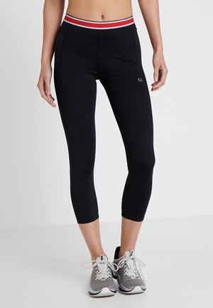 Schwarze, eng anliegende, kurz geschnittene Leggings mit einem Taillenbund in Rot, Weiß und Schwarz gestreift, kleinem "Calvin Klein"-Logo, getragen mit grauen und weißen Sportschuhen.