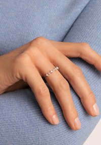 Anello in argento con piccole pietre trasparenti incastonate in un delicato motivo, indossato su una mano contro uno sfondo di tessuto azzurro chiaro texturizzato.