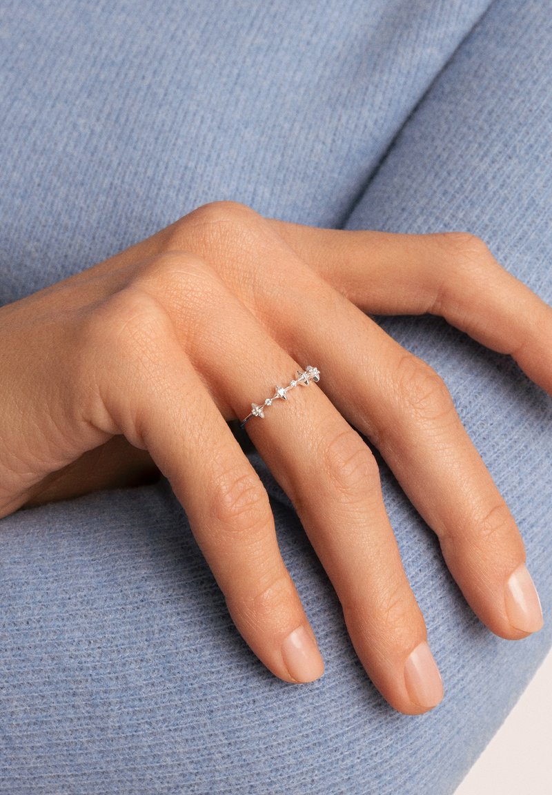 Anello in argento con piccole pietre trasparenti incastonate in un delicato motivo, indossato su una mano contro uno sfondo di tessuto azzurro chiaro texturizzato.