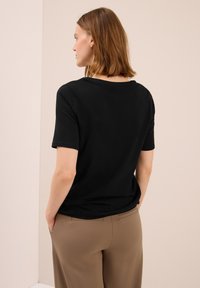 Eine Frau mit schulterlangen Haaren trägt ein schwarzes T-Shirt mit kurzen Ärmeln und beige Hosen, steht mit den Händen in den Taschen und schaut weg.