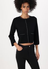 Cárdigan corto negro con escote redondo, adornado con tachuelas plateadas en la parte frontal. Combinado con pantalones negros de talle alto con cremallera.