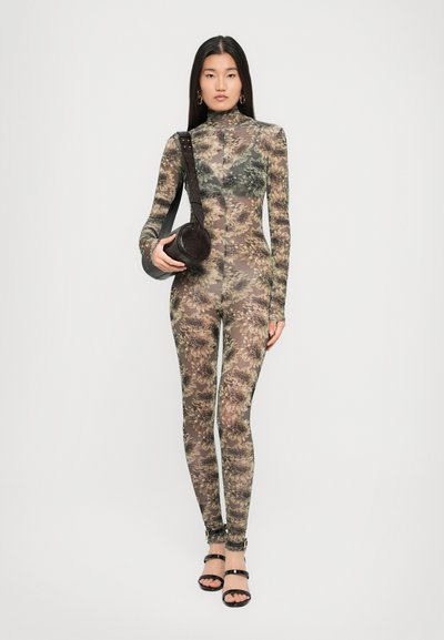 Mujer vestida con un mono de cuerpo entero, transparente y de estampado floral, sosteniendo un bolso redondo negro y usando tacones negros con tiras.
