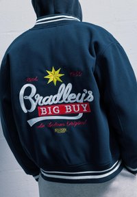 Námořnická modrá bunda s bílými pruhovanými manžetami, se barevným vyšitým textem: "Bradley's BIG BUY," založeno 1960. Text obsahuje žlutou hvězdnou emblému.