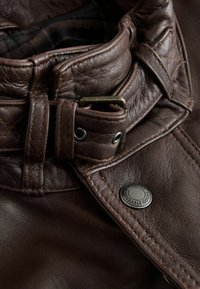 Braune Lederjacke mit Kragen und gestepptem Muster, metallischem Schnallenverschluss und Druckknopfdetails. Das Innenfutter hat ein dunkles Karomuster.