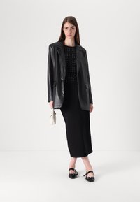 Blazer oversize in pelle nera sopra un top nero con borchie argentate, abbinato a una lunga gonna nera e scarpe con tacco nere. Piccola borsa bianca.