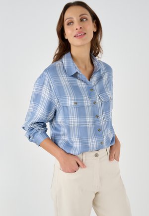 Camicia a scacchi blu con strisce azzurre e bianche, dotata di due tasche sul petto e maniche arrotolate, abbinata a pantaloni beige.