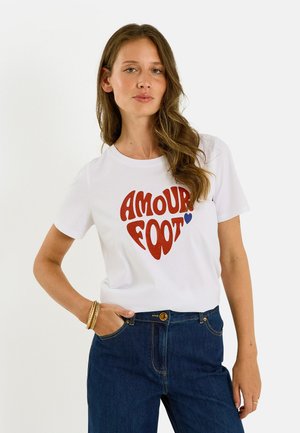 Jeune femme aux longs cheveux ondulés portant un t-shirt blanc avec le texte rouge "AMOUR FOOT" et un jean bleu, debout avec une main dans la poche.