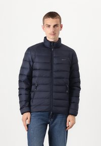 Donkerblauwe gewatteerde jas met hoge kraag, ritssluiting en twee zijzakken. Kenmerkt zich door een horizontaal gestikt patroon en een subtiele merklogo.