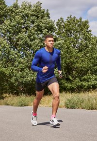 Maglietta sportiva blu a maniche lunghe con strisce nere, abbinata a pantaloni corti neri e scarpe da tennis bianche con dettagli rosa. Corre su un sentiero.