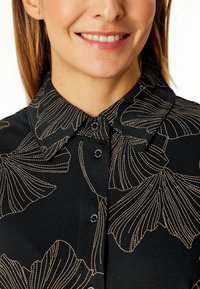 Black shirt with floral patterns in gold and beige. Features a collared neck and button closure. Smooth texture with a tailored fit.