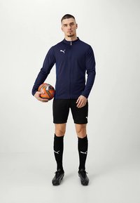 Man in navy jacket and black soccer shorts holds an orange soccer ball, wearing black knee-high socks and cleats, standing against a plain background.