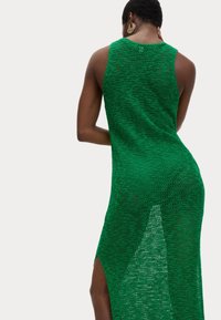 Grünes Maxi-Kleid aus Strick mit ärmellosem Design, ausgestattet mit einem seitlichen Schlitz und offener Maschenstruktur. Auffällige Rückendetails umfassen einen geschwungenen Ausschnitt.