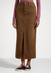 Brun denim maxikjol med en framslits, strukturerad design och kontrasterande sömmar, kombinerad med bruna sneakers och en rosa långärmad topp.