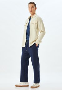 Chaqueta texturizada beige sobre una camiseta azul, combinada con pantalones azul marino y zapatillas blancas con suela marrón. Dos bolsillos en el pecho de la chaqueta.
