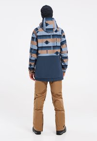 Veste de ski avec un motif géométrique bleu et beige, section inférieure marine, poignets élastiques et capuche ajustable. Associée à un pantalon de neige beige.