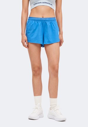 Nő kék sport rövidnadrágban, fehér Under Armour crop topban, fehér zokniban és fehér tornacipőben, sima háttér előtt áll.