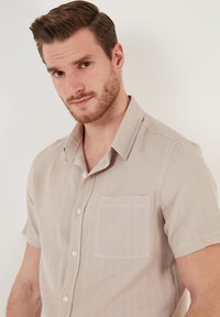 Camisa de botones de manga corta, color beige claro, con un bolsillo en el pecho, tejido texturizado y un diseño de cuello relajado.