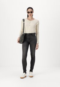 Kremfarget ribbet topp med lange ermer og knapper, svarte høytalende skinny jeans, grå crossover-veske, og hvite joggesko med gummisåler.
