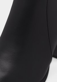 Κοντινό πλάνο μαύρου δερμάτινου μποτάκι με ομαλή υφή, καθαρές ραφές και χοντρό τακούνι σε λευκό φόντο.