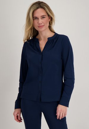 Blonde vrouw met gouden creolen en een marineblauwe blouse met lange mouwen en knopen, gecombineerd met bijpassende broek, staand tegen een neutrale achtergrond.
