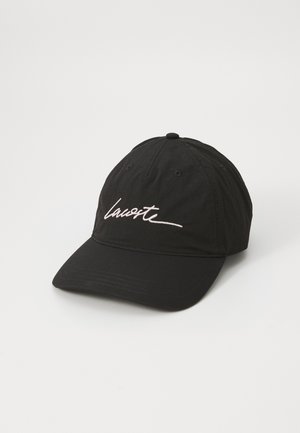 Juodas baseball kepurė, pagaminta iš audinio, su lenktu kraštu. Priekyje yra balta siuvinėta "Lacoste" logo. Galinėje dalyje yra reguliuojamas dirželis.