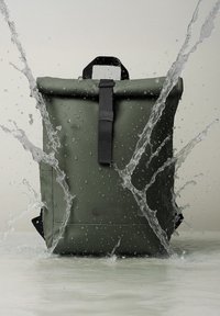 Zaino roll-top verde resistente all'acqua con una cinghia nera e accessori, caratterizzato da un tessuto testurizzato e schizzi d'acqua intorno.