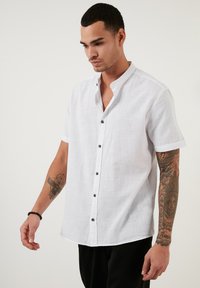 Cămașă albă din in cu mâneci scurte, cu guler tip bandă, nasturi negri și o țesătură texturată. Asortată cu pantaloni casual negri.