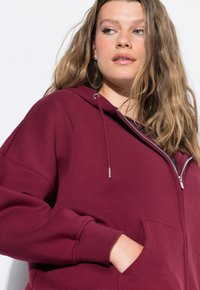 Sudadera burdeos hecha de tela suave, con cierre de cremallera, cordones ajustables y bolsillos laterales, además de puños y dobladillo acanalados.