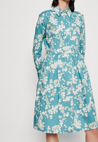 En turkos bomulls klänning med ett blommönster i vitt och kräm. Den har en knäppning fram, en krage och ett samlat midja.