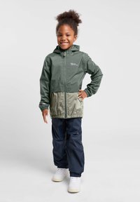 Grüne und khakifarbene Regenjacke mit Kapuze, elastischen Bündchen und Reißverschluss, kombiniert mit marineblauen wasserdichten Hosen und weißen Sneakers.