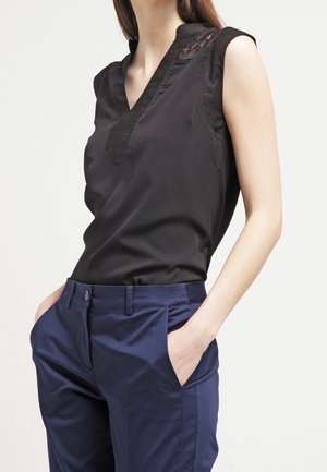 Czarna bluzka bez rękawów z koronkową lamówką na ramionach i dekolcie, w połączeniu z granatowymi, dopasowanymi spodniami z wysokim stanem, ręce w kieszeniach.