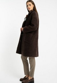 Brącoat, teksturowany płaszcz z wysokim kołnierzem, zapięciem na guziki oraz szarym akcentem w formie kwiatu. Nosi się go na wzorzyste spodnie i brązowe buty.