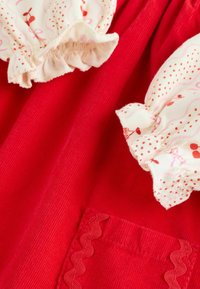 Red corduroy fabric with a small patch pocket and cream-colored ruffled cuffs featuring red and pink floral patterns.