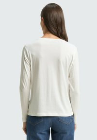 Vrouw met lang bruin haar draagt een witte blouse met lange mouwen en een spijkerbroek, staande met haar rug naar de camera tegen een effen lichtgrijze achtergrond.