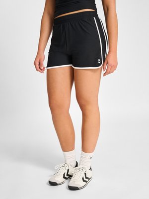 Kvinde iført sorte atletikshorts med hvid kant, hvide sokker og hvide sneakers med sorte chevron-detaljer mod en ensartet lys baggrund.