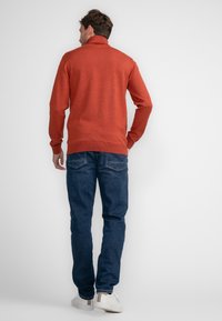 Oranger gestrickter Pullover mit hohem Kragen, kombiniert mit blauen Jeans. Weiße Turnschuhe mit beige Akzent an der Ferse.
