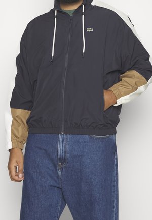 Man draagt een zwart jack met rits, met mouwen in blokkleuren van beige en wit, en blauwe spijkerbroek. Hij staat met één hand in zijn zak.