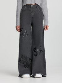 Jeans neri a vita alta e gambe larghe con toppe a forma di stella e la scritta "Born Rocky", orli sfrangiati, indossati con sneaker bianche e maglione grigio corto.