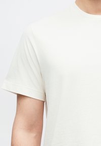 Světle béžové bavlněné tričko s kulatým výstřihem a krátkými rukávy. Tkanina působí měkce se hladkou texturou a jednoduchým designem.