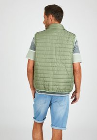 Grüne, flauschige Weste mit einer gesteppten Textur, ärmellosem Design und hohem Kragen, getragen über einem gestreiften Hemd und kombiniert mit hochgekrempelten Jeansshorts.