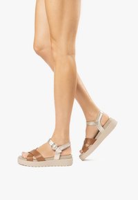 Sandali strappy marroni e metallici con una base ampia, cinturino alla caviglia regolabile e suola testurizzata. Presentano un accento circolare metallico.