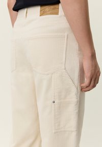 Krem hlače od korduroga s vertikalnim rebrastim uzorkom, bočnim teretnim džepom, stražnjim džepovima i smeđom kožnom oznakom na pojasu.