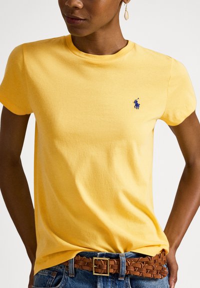 Ženska, oblečena v rumeno majico s kratkimi rokavi z modrim logotipom, modre kavbojke in rjav prepleten pas, videti je viseča uhani.