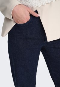 Main avec des ongles rose très clair dans la poche avant d'un jean bleu foncé, portant une veste beige sur un top en dentelle blanche.