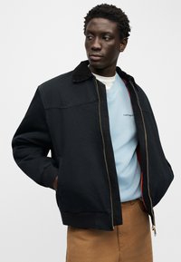 Νεαρός άνδρας που φοράει μαύρο μπουφάν πάνω από ανοιχτό μπλε πουλόβερ και καφέ παντελόνι, κοιτάζοντας προς τα αριστερά με τα χέρια στις τσέπες του μπουφάν.