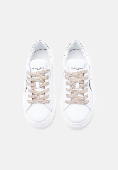 Λευκά δερμάτινα sneakers με μπεζ κορδόνια, στρογγυλή μύτη και λαστιχένια σόλα. Διαθέτουν διακριτικό λογότυπο στη γλώσσα και μινιμαλιστικά σχεδιαστικά στοιχεία.