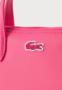 Sac fourre-tout synthétique rose avec une surface texturée, arborant un logo Lacoste blanc brodé à l'avant et une sangle assortie rose.