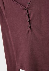 Long-sleeve burgundy top made of ribbed fabric featuring a buttoned placket with black buttons along one side and a rounded hem.