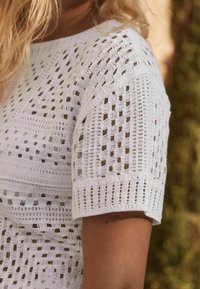 Top de punto de ganchillo blanco con un patrón texturizado y diseños de calados, mangas cortas y dobladillo recto.