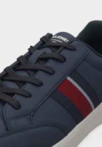 Ναυτικό μπλε αθλητικό παπούτσι με μαύρα κορδόνια, πλευρικές ρίγες σε κόκκινο, λευκό και ναυτικό μπλε, και λευκή σόλα, με το λογότυπο Jack & Jones στην γλώσσα του παπουτσιού.